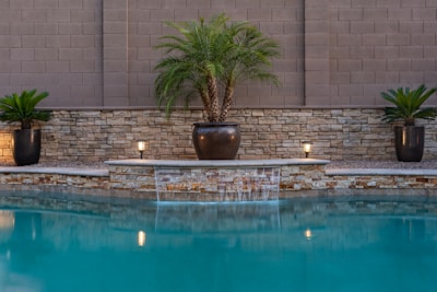 A serene pool area with blue water in the foreground, flanked by a stone wall and neatly arranged potted plants. The plants are set in black pots, and two small lights are visible, adding a subtle illumination. A gentle waterfall feature is present in the center, enhancing the tranquility of the setting.