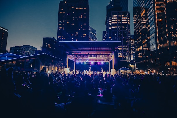 A public outdoor concert is taking place in a city setting during the evening. The stage is lit with colorful lights, and a large crowd of people are gathered in the foreground, sitting on the grass and enjoying the performance. Surrounding the scene are tall buildings with illuminated windows, creating an urban backdrop.