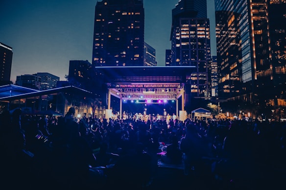 A public outdoor concert is taking place in a city setting during the evening. The stage is lit with colorful lights, and a large crowd of people are gathered in the foreground, sitting on the grass and enjoying the performance. Surrounding the scene are tall buildings with illuminated windows, creating an urban backdrop.