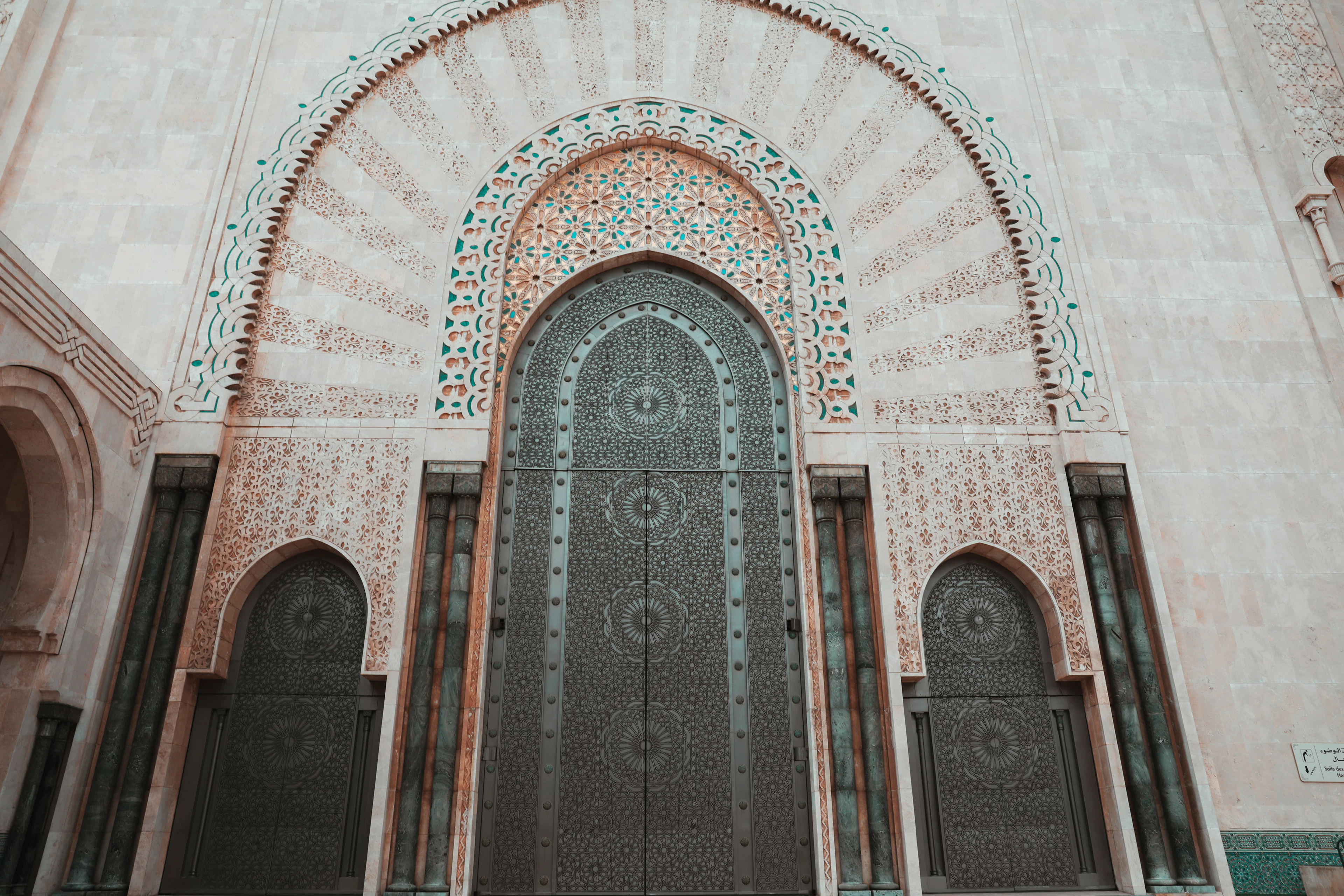 An ornate pink stone archway with green mosaic tilework frames a pair of dark studded doors, in Casablanca's Habous quarter.