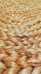 A close-up view of tightly woven natural fiber, likely jute or sisal, showcasing a braided pattern. The texture appears rough and tactile, with a natural sheen reflecting light.
