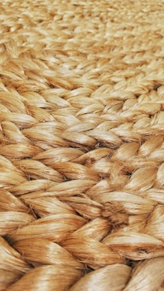 A close-up view of tightly woven natural fiber, likely jute or sisal, showcasing a braided pattern. The texture appears rough and tactile, with a natural sheen reflecting light.