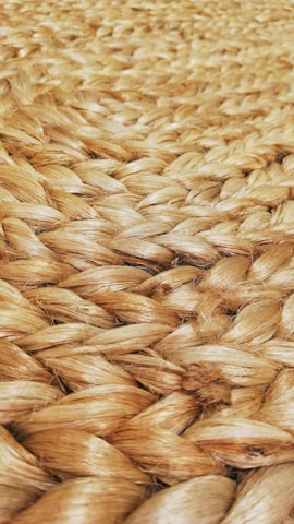 A close-up view of tightly woven natural fiber, likely jute or sisal, showcasing a braided pattern. The texture appears rough and tactile, with a natural sheen reflecting light.