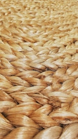 A close-up view of tightly woven natural fiber, likely jute or sisal, showcasing a braided pattern. The texture appears rough and tactile, with a natural sheen reflecting light.