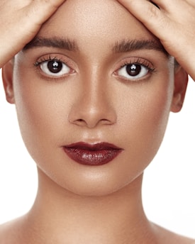 A close-up portrait of a person with well-defined facial features, focusing on their eyes and lips. The hands are gently placed on the forehead, and the makeup is minimal yet striking with bold lipstick.