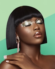 A person with smooth, dark skin and short, straight black hair adorned with shimmering makeup and embellishments around their eyes. The eyes are closed, showcasing sparkly, silver-green eyeshadow and small shining pearls around the eyelids. The lips are glossy and appear in a natural shade, while the person wears large, elegant earrings with a combination of sparkling elements and pink tassels. The background features a vibrant mint green diagonal split.