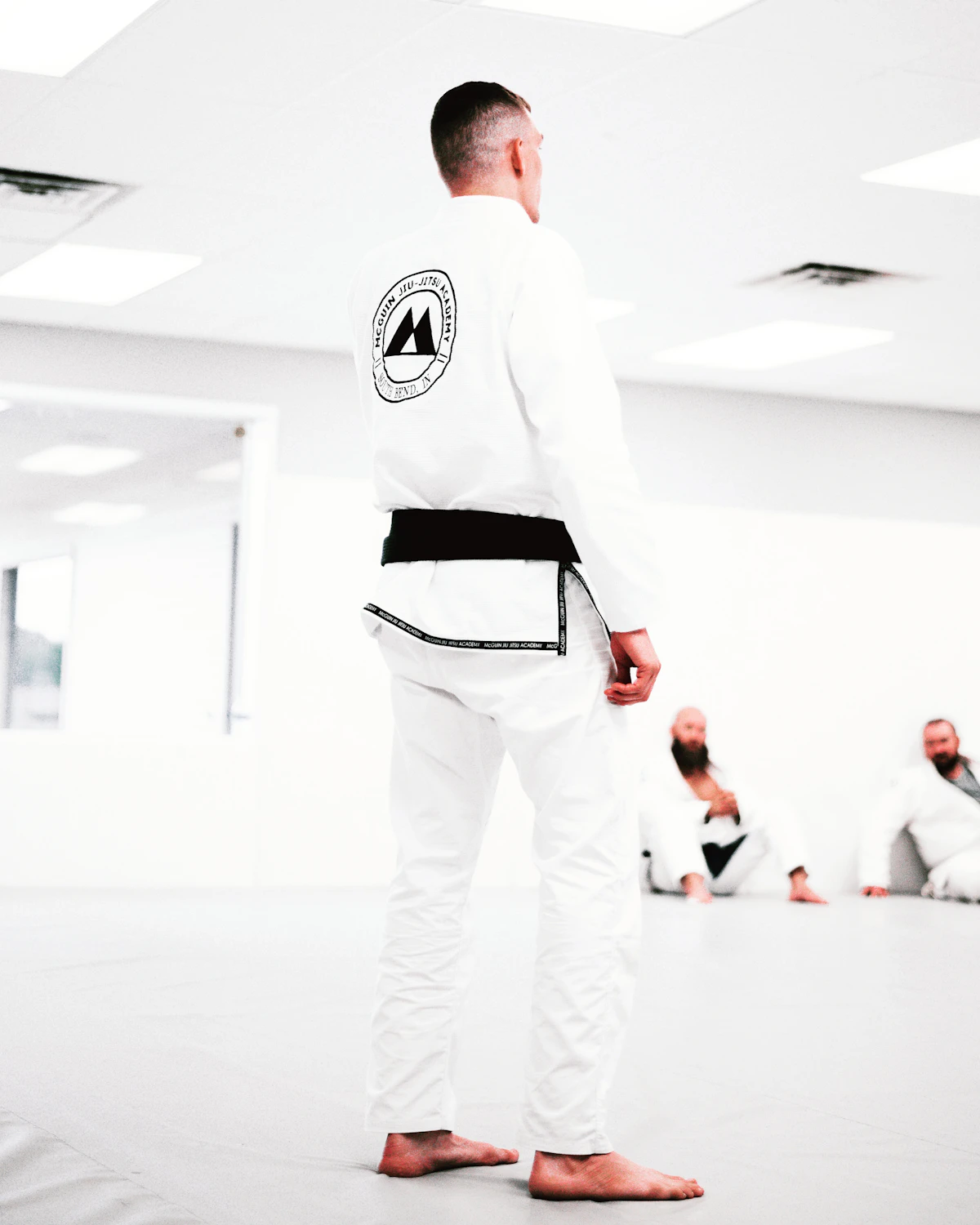 A black belt in a white gi, seen from behind on the mats of a bright academy