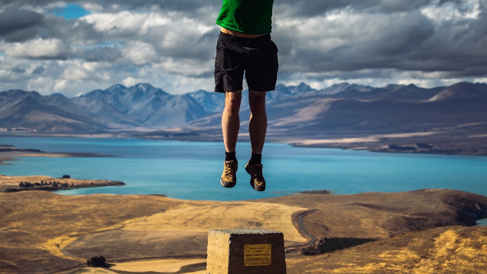 man in green shirt and black shorts standing on brown rock near blue sea during daytime