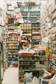 A small, densely packed grocery store filled with shelves displaying a variety of dry goods, beverages, and snacks. The shelves are stacked to the ceiling with products including canned goods, bottles, and packets. A person, likely a store clerk, stands behind a counter surrounded by assorted snacks and food items. The surroundings are colorful and visually busy with lots of packaging and branding visible.