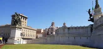 A monument with elaborate sculptures is set against a bright blue sky. The architecture features grand buildings in a classical style, with ornate domes and historic structures. A well-maintained grassy area is in the foreground, and an Italian flag is visible on one of the sculptures.