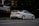 A white sports car is parked on a quiet street next to a building with a sign in Cyrillic script. The car has a prominent spoiler, sleek design, and sports wheels. The surrounding environment is calm with trees, grass, and a dimly lit establishment with lights visible inside.