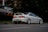 A white sports car is parked on a quiet street next to a building with a sign in Cyrillic script. The car has a prominent spoiler, sleek design, and sports wheels. The surrounding environment is calm with trees, grass, and a dimly lit establishment with lights visible inside.