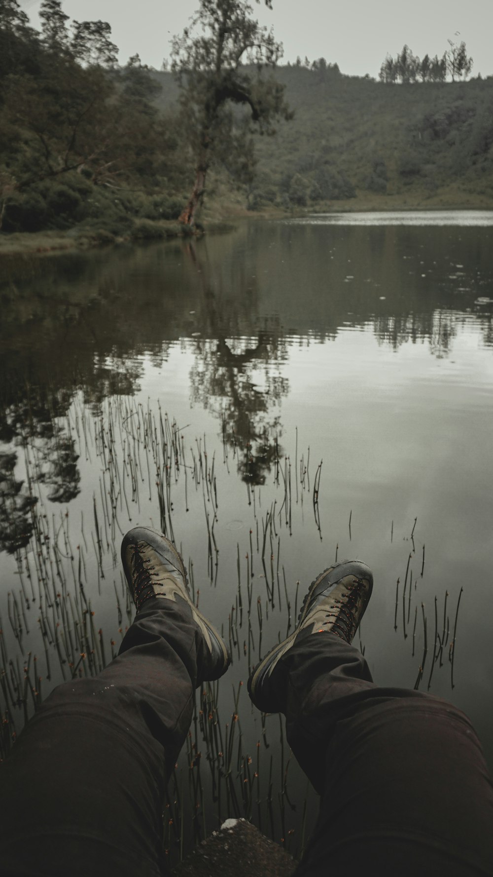 Persona con pantalones marrones y zapatos negros sentada en una roca cerca del lago durante el día