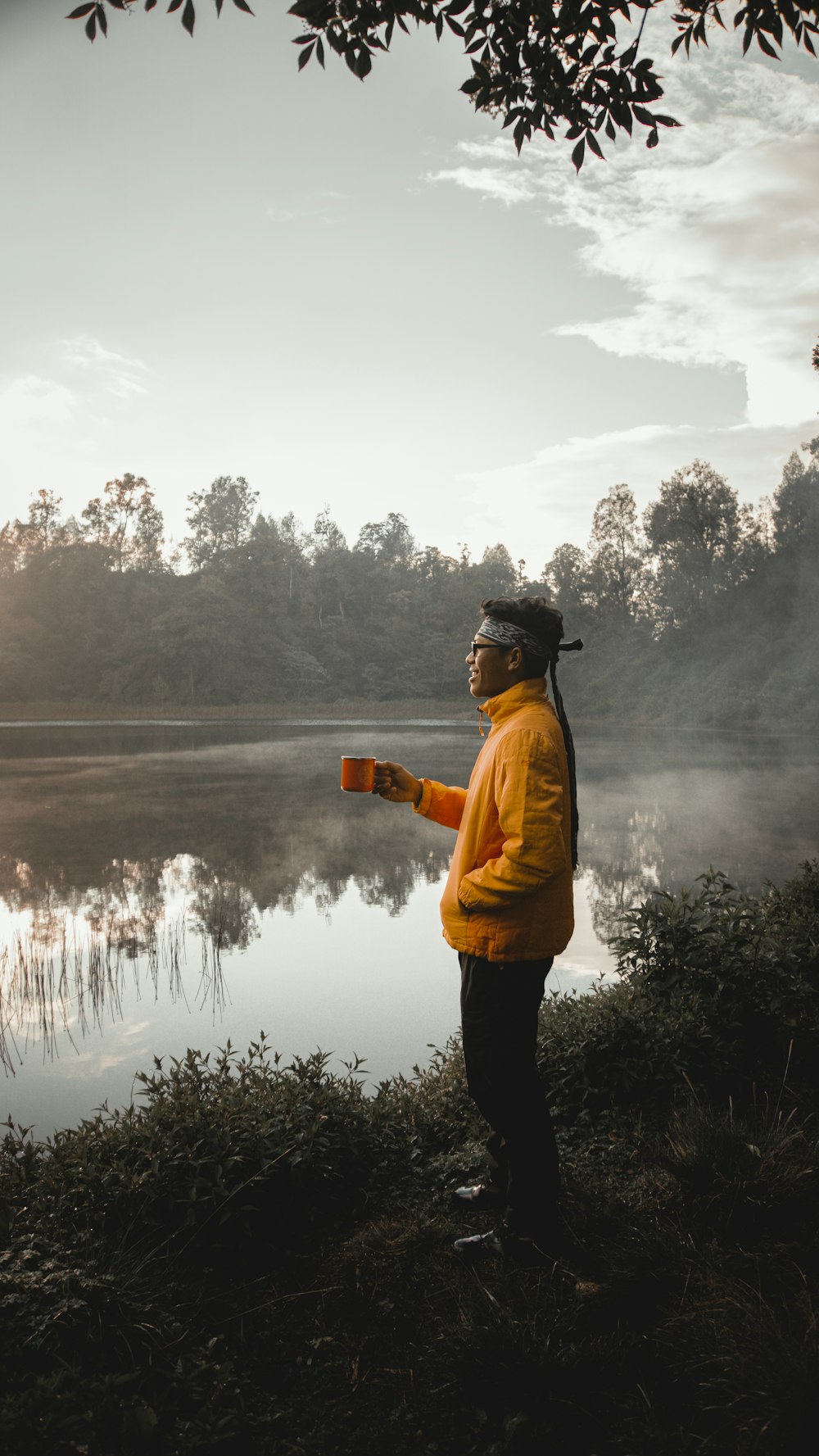 man in yellow jacket and black pants holding brown cup standing on green grass near lake