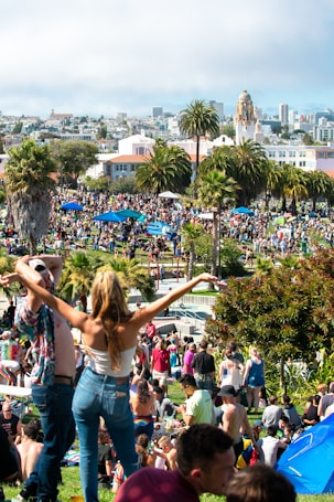 A lively park scene full of people enjoying a sunny day. Many individuals are lounging on the grass or walking around, with some carrying colorful umbrellas. Palm trees and various green plants are scattered throughout the area. In the background, there are buildings and a distant city skyline. The atmosphere is vibrant and bustling.