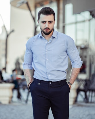 A man stands confidently outdoors, wearing a light blue long-sleeve shirt and navy pants. His hands are in his pockets and he has a groomed beard. The background shows an urban setting with blurred architectural elements and people.