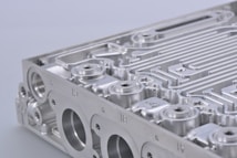 A close-up view of a metallic component, featuring intricate grooves and perfectly machined surfaces. The object appears to be part of an industrial or mechanical structure with multiple circular openings and linear patterns.