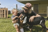 A young man with long hair, wearing sunglasses and a cap, crouches on the grass beside a toddler and a small black and brown goat. The toddler appears to be hesitant or startled by the goat while the man is gently guiding the toddler's interaction. The background includes a fenced area and a building, suggesting a rural or farm setting.