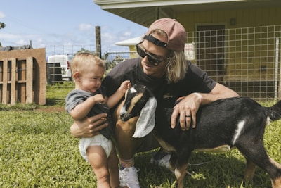 A young man with long hair, wearing sunglasses and a cap, crouches on the grass beside a toddler and a small black and brown goat. The toddler appears to be hesitant or startled by the goat while the man is gently guiding the toddler's interaction. The background includes a fenced area and a building, suggesting a rural or farm setting.
