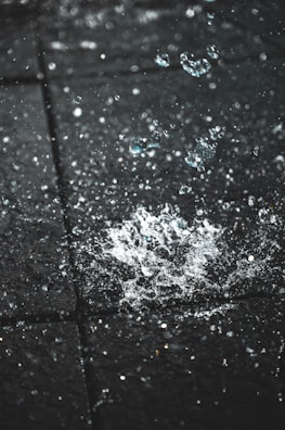 Water droplets are captured in mid-air as they splash upon hitting a dark textured surface. The surface is made up of square tiles, and the light highlights the clarity and motion of the water.