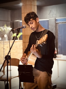 A person is playing an acoustic guitar, standing in front of a microphone, and appears to be focused on music. The background includes a brick pillar, some indoor plants, and a table with a drink bottle. The setting seems casual and relaxed, possibly a small venue or caf&eacute;.