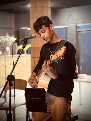 A person is playing an acoustic guitar, standing in front of a microphone, and appears to be focused on music. The background includes a brick pillar, some indoor plants, and a table with a drink bottle. The setting seems casual and relaxed, possibly a small venue or café.