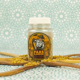 A jar of honey with a vintage-style label depicting a beekeeper is surrounded by wooden spoons filled with bee pollen. The background features a repetitive geometric pattern in light blue and white.