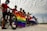 A group of cyclists stands outdoors, holding bicycles and two flags: a rainbow flag and a flag with a single star. The cyclists are wearing helmets and masks, suggesting a focus on safety. The setting is open with a cloudy sky overhead.
