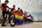 A group of cyclists stands outdoors, holding bicycles and two flags: a rainbow flag and a flag with a single star. The cyclists are wearing helmets and masks, suggesting a focus on safety. The setting is open with a cloudy sky overhead.