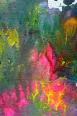 A vibrant and abstract watercolor painting featuring a mix of colors such as green, pink, yellow, and blue. The colors blend and flow into each other, creating an effect reminiscent of foliage or flowers. The image has a textured, almost fabric-like appearance with irregular shapes and vivid patches of color.