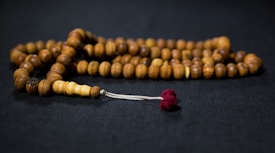 A string of wooden prayer beads is arranged on a dark surface. The beads are smooth and rounded, ranging in shades of brown, with a small red tassel at one end.