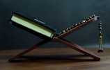 A sacred book with a black cover rests open on a wooden book stand, accompanied by a black and gold prayer beads. The background is dark and minimalistic, emphasizing the objects in the foreground.