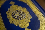 A close-up view of a textured book cover featuring intricate gold Arabic calligraphy set against a deep blue background. The ornate design includes geometric patterns and floral motifs, enhancing its elegant appearance.