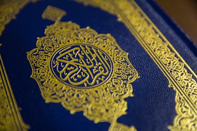 A close-up view of a textured book cover featuring intricate gold Arabic calligraphy set against a deep blue background. The ornate design includes geometric patterns and floral motifs, enhancing its elegant appearance.