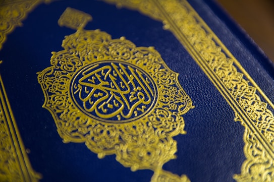 A close-up view of a textured book cover featuring intricate gold Arabic calligraphy set against a deep blue background. The ornate design includes geometric patterns and floral motifs, enhancing its elegant appearance.