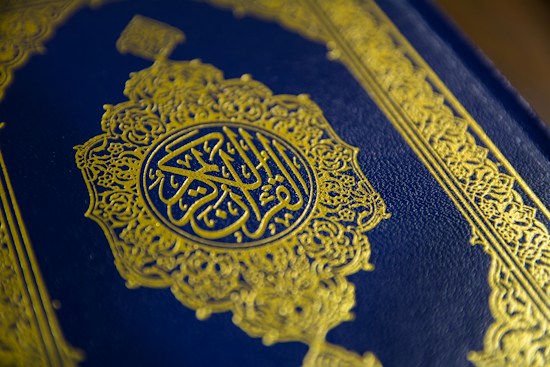 A close-up view of a textured book cover featuring intricate gold Arabic calligraphy set against a deep blue background. The ornate design includes geometric patterns and floral motifs, enhancing its elegant appearance.