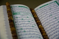 An open book with Arabic script is displayed, likely a religious text. A string of brown prayer beads is placed across the pages.