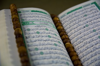 An open book with Arabic script is displayed, likely a religious text. A string of brown prayer beads is placed across the pages.