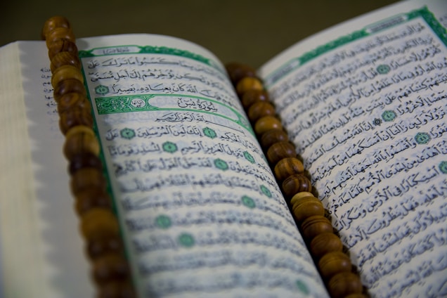 An open book with Arabic script is displayed, likely a religious text. A string of brown prayer beads is placed across the pages.