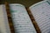 An open book with Arabic script is displayed, likely a religious text. A string of brown prayer beads is placed across the pages.