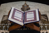 A beautifully decorated book with Arabic script open on a wooden stand. The backdrop features a richly patterned, fabric depicting a building with ornate architecture. The book's pages are adorned with intricate geometric borders.
