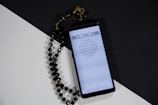 A smartphone displays a page of Arabic text probably from a religious script. Next to it, black prayer beads with gold accents are arranged on a surface divided into black and white sections, creating a contrasting background.