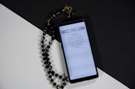 A smartphone displays a page of Arabic text probably from a religious script. Next to it, black prayer beads with gold accents are arranged on a surface divided into black and white sections, creating a contrasting background.