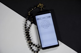 A smartphone displays a page of Arabic text probably from a religious script. Next to it, black prayer beads with gold accents are arranged on a surface divided into black and white sections, creating a contrasting background.