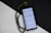 A smartphone displays a page of Arabic text probably from a religious script. Next to it, black prayer beads with gold accents are arranged on a surface divided into black and white sections, creating a contrasting background.