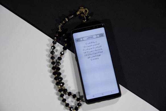 A smartphone displays a page of Arabic text probably from a religious script. Next to it, black prayer beads with gold accents are arranged on a surface divided into black and white sections, creating a contrasting background.