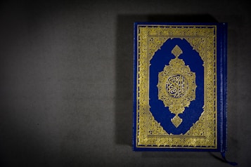 A beautifully designed book with a blue cover and intricate gold patterns featuring Arabic script in the center. The book is placed on a dark gray surface, with a tassel visible at the bottom.