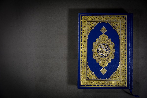A beautifully designed book with a blue cover and intricate gold patterns featuring Arabic script in the center. The book is placed on a dark gray surface, with a tassel visible at the bottom.