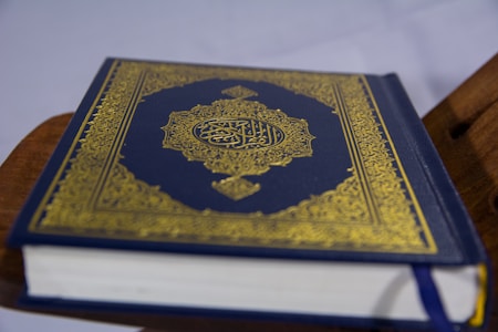 A closed book with an ornate cover featuring intricate gold patterns and Arabic script, resting on a wooden stand. The book appears to be a religious text, possibly the Quran, with a dark blue cover.