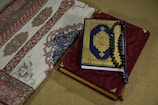 A Quran with a blue and gold ornate cover rests on top of a red prayer rug adorned with intricate embroidery. Beside it, a section of an intricately designed fabric with floral and geometric patterns is visible.